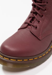 Stivaletto alla caviglia in pelle bordeaux con punta tonda, lacci piatti e cuciture gialle lungo la suola spessa. Dettaglio della superficie leggermente strutturata.