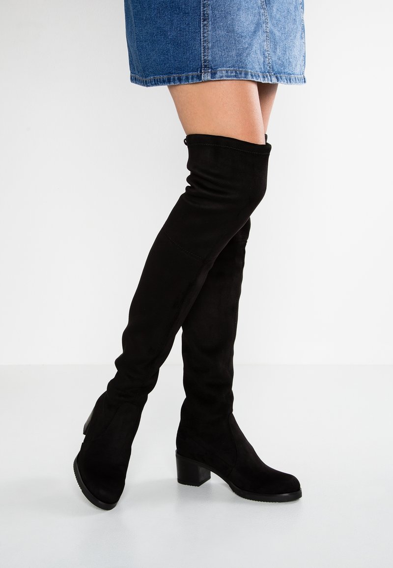 Jambes portant une jupe en jean bleue et des bottes noires en suède montant au-dessus du genou avec des talons carrés, debout sur un sol blanc sur un fond uni.