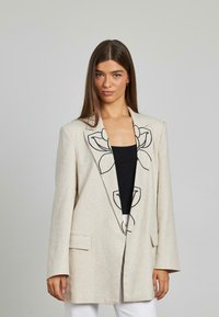 Blazer in lino beige con ricami floreali neri sulla parte frontale, con taglio su misura, revers a punta e due tasche anteriori.
