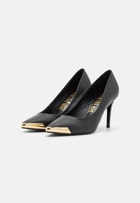 Escarpins noirs à bout pointu en cuir lisse, présentant un bout métallique doré et un talon fin. L'intérieur comporte un imprimé logo.