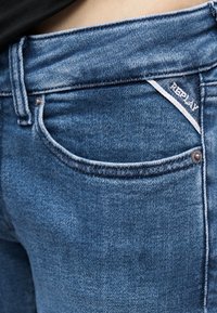 Εγγύς πλάνο τσεπάκι από μπλε τζιν με εμφανή ετικέτα της μάρκας Replay και λεπτομέρεια από μεταλλικό στήριγμα σε μαύρο φόντο μέσης.