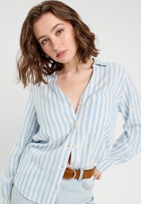 Chemise légère à boutons, rayée bleue et blanche, avec une poche avant. Texture douce et coupe décontractée, assortie à une ceinture marron.