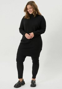 Črna pletena obleka s visokim ovratnikom, združena s slim-fit črnimi hlačami in črnimi debelimi loaferji, prikazana na nevtralni podlagi.
