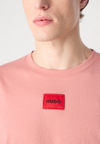 Világos rózsaszín pamut póló, piros téglalap alakú folttal, rajta fekete betűkkel "HUGO" felirattal. Kerek nyakkivágású és rövid ujjú. Sima tapintású.