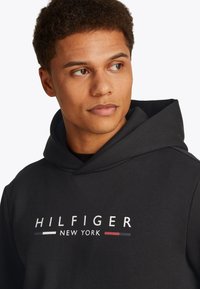 Young man with short curly hair wearing a black hoodie with "HILFIGER NEW YORK" text, looking to the right on a plain background.