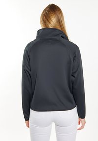 Svart pulloverjacka med hög krage, tillverkad av strukturerat tyg. Har raglanärmar och platta sömmar. Sömmen slutar ovanför höfterna.