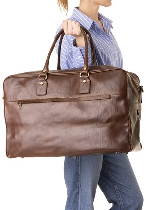 Donna con camicia a righe blu e jeans che tiene una grande borsa da viaggio in pelle marrone per le maniglie di lato.
