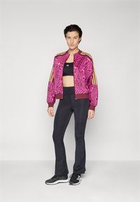 Roze geblokte bomberjack met bordeauxrode afwerking, ritssluiting en gouden strepen. Zwarte crop top en wijde zwarte broek, gecombineerd met zwarte sneakers.
