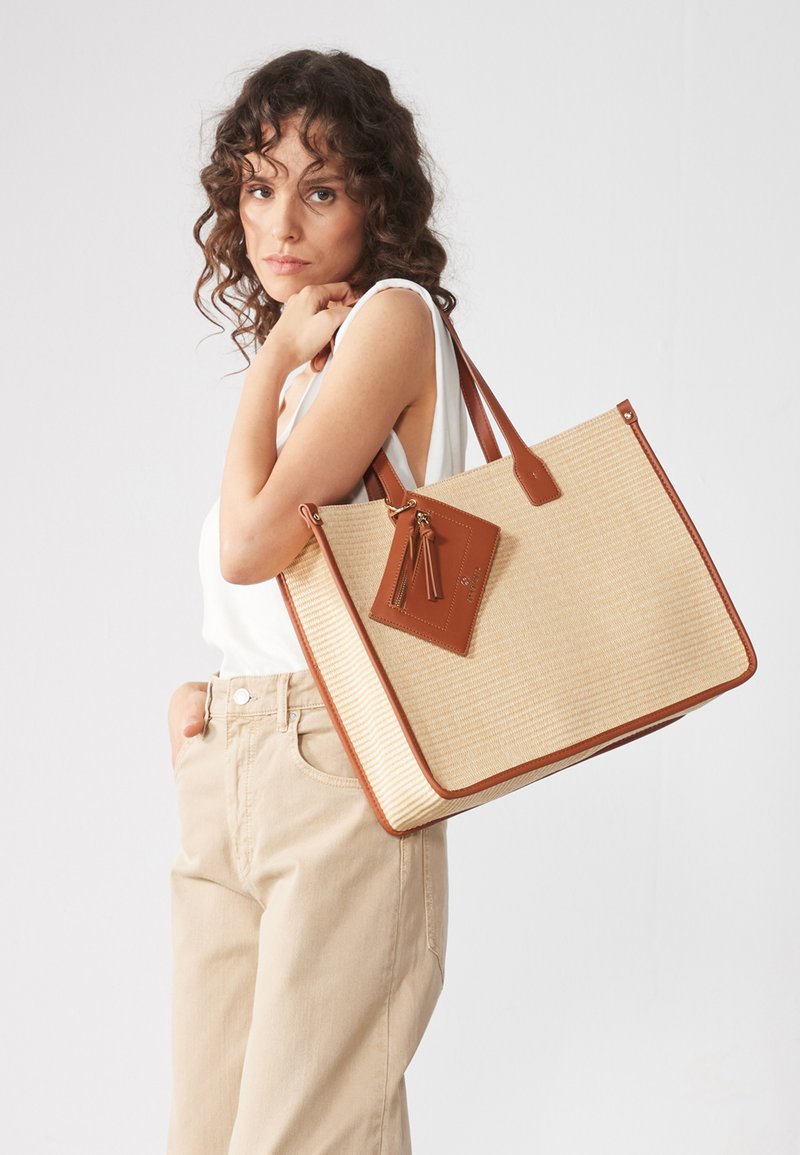 Borsa tote beige con tessuto testurizzato e dettagli in pelle marrone. Presenta manici lunghi e un'etichetta removibile con frange in pelle.