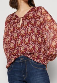 Donna che indossa una blusa bordeaux trasparente a maniche lunghe con un motivo geometrico astratto, infilata in jeans di denim blu scuro.