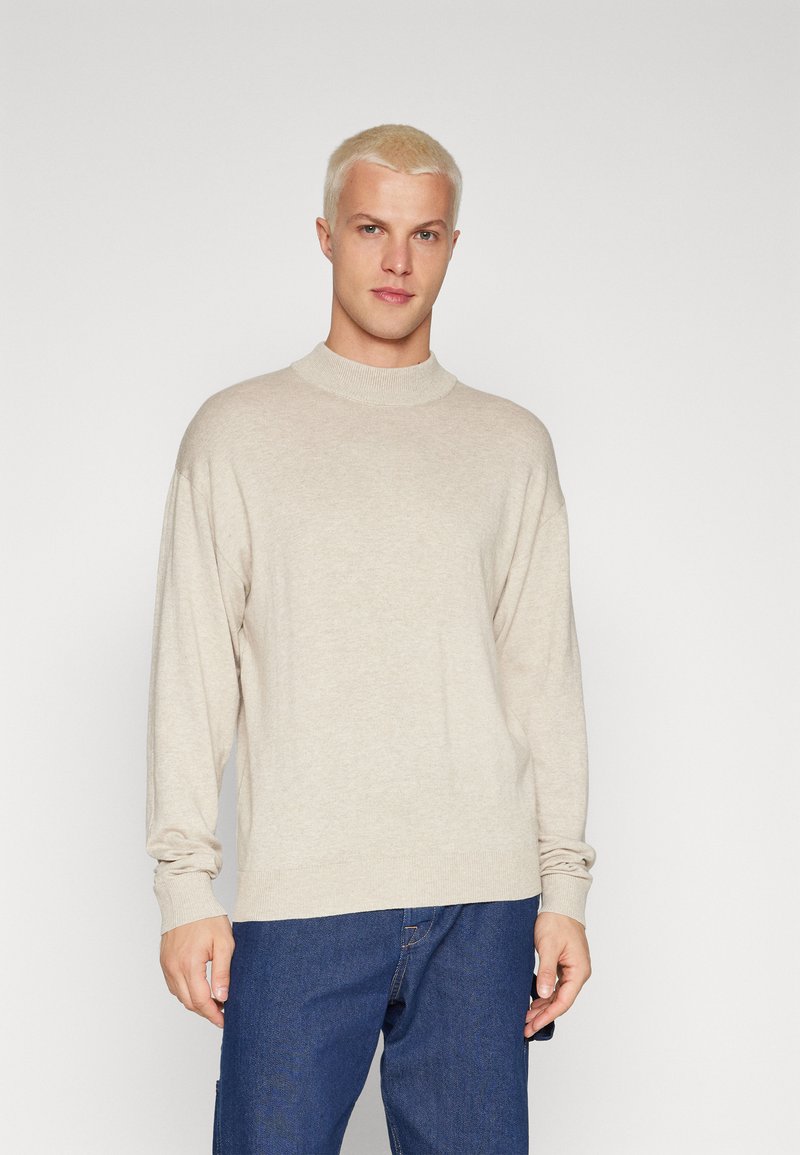 Suéter de punto de color beige claro con cuello alto, mangas largas y un corte holgado. Presenta una textura suave y puños y bajo acanalados.