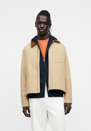 Mladi moški s kitkami, oblečen v bež jakno čez mornarsko moder pulover z zadrgo in oranžno majico, kombinirano z belimi hlačami, na belem ozadju.