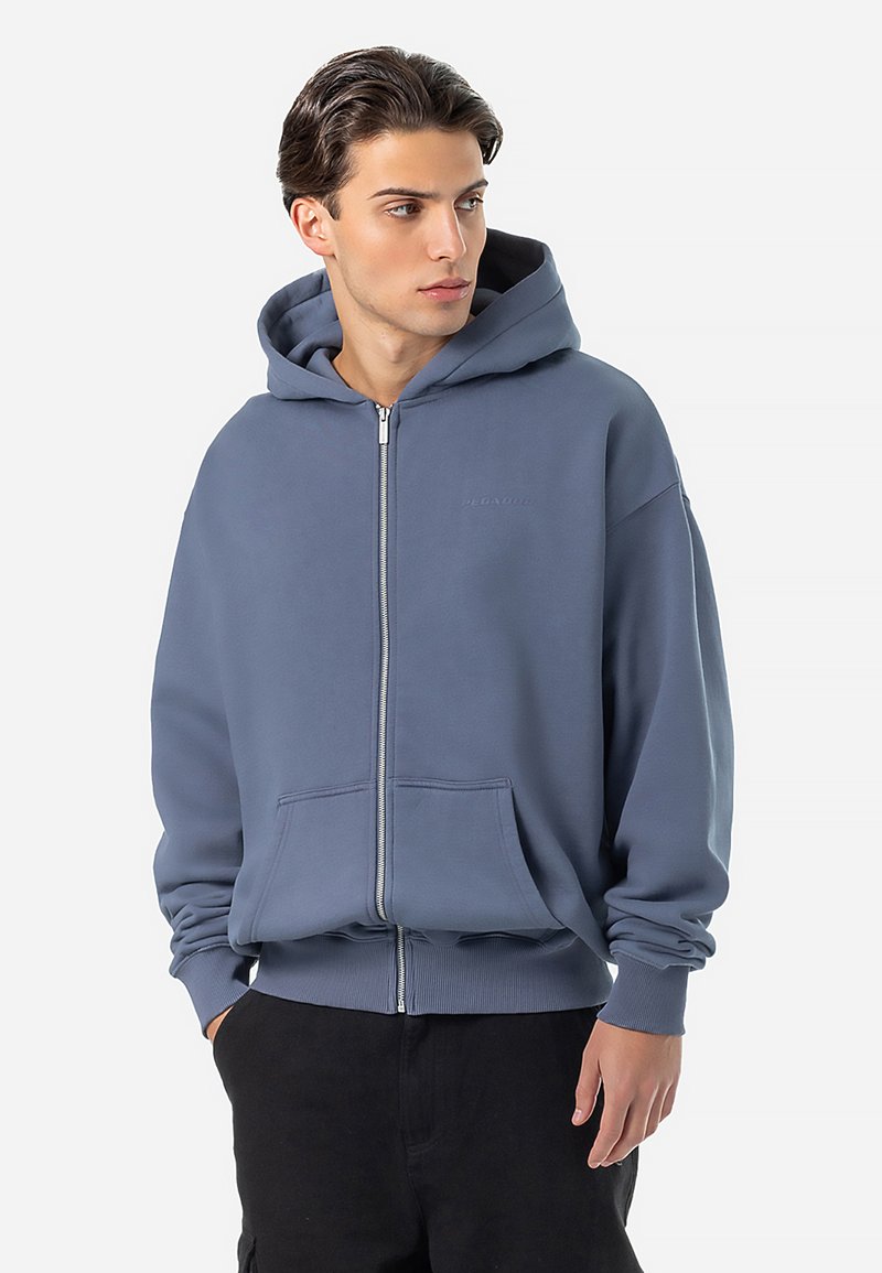 Mikina s kapucňou na zips v mäkkej, tlmenej modrej látke. Obsahuje kengurské vrecká, rebrované manžety a decentné logo značky na hrudi.