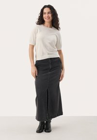 Un pull à manches courtes gris clair associé à une jupe maxi en denim noire avec une fente à l'avant et des bottines noires, se tenant sur un fond neutre.