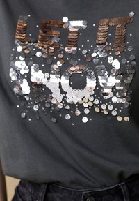 Dark gray t-shirt featuring the text "LET IT NOW" made of circular sequin accents in silver and bronze, creating a shimmering effect.