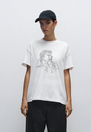 Hvid bomulds t-shirt med en sort stregtegning af en figur, med løs pasform og korte ærmer. Båret med en sort kasket.