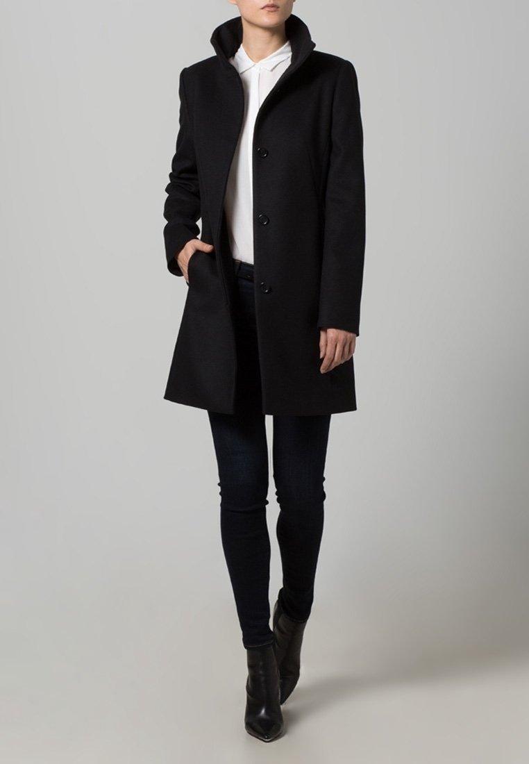 Femme portant un manteau noir boutonné sur un sweat à capuche blanc, un jean skinny foncé et des bottines noires, debout avec une main dans la poche.