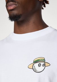 T-shirt bianca in cotone con una grafica raffigurante una figura rotonda in stile cartone animato, con un cappello a tesa larga e occhi a spirale sul petto.