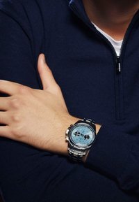 Sidabrinis nerūdijančio plieno laikrodis su mėlynu ciferblatu, turintis du juodus subciferblatus ir sidabrinius akcentus, dėvimas ant rankos prieš tamsią megztinę.