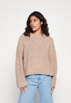 Pull en tricot texturé beige clair avec des éclats pastel, col rond, épaules tombantes et bord inférieur court, associé à un jean bleu clair.