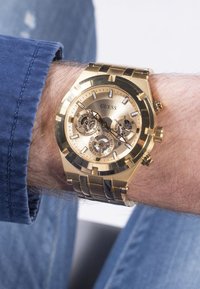 Złoty zegarek Guess o okrągłej tarczy, funkcjami chronografu i metalową bransoletą. Posiada trzy małe tarcze oraz teksturowane wykończenie bezel.