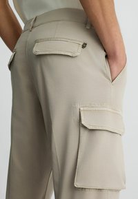 Beige cargo nadrág sima szövet textúrával, két hátsó zsebbel fedéllel és oldalsó zsebekkel a fokozott funkcionalitás érdekében.