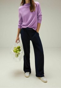 Ljuslila crew-neck tröja, mörkblå vida byxor, vita sneakers, håller en genomskinlig väska med gröna äpplen. Minimalistisk design.