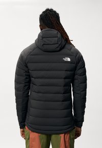 Giacca a vento in nero uniforme, presenta un cappuccio e un design trapuntato con sezioni orizzontali. Logo nella parte superiore sinistra. Tessuto liscio.
