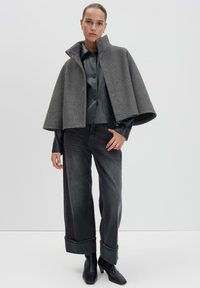 Grau wollener Umhang mit hohem Kragen und Druckknopfverschluss, kombiniert mit einem schwarzen, knopfbaren Hemd aus Kunstleder und weiten, dunklen Jeanshosen.