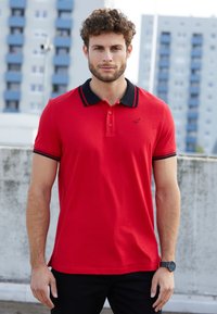 Rotes Poloshirt mit einem schwarzen Kragen und Bündchen, das ein Logo auf der Brust zeigt, kurze Ärmel und einen geraden Saum hat. Getragen mit dunklen Hosen.