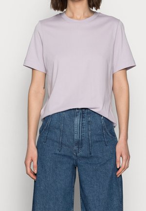 Kvinde iført en ensfarvet lyse lilla kortærmet t-shirt, som er stoppet ned i højtaljede, løstsiddende blå jeans.