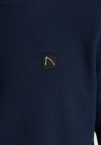 Pull bleu marine texturé avec un motif en maille côtelée et un petit patch noir arborant un motif géométrique blanc sur la poitrine.