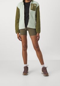 Persona che indossa un giacca in fleece verde bicolore, pantaloni corti da bicicletta oliva, calze bianche e scarpe da escursionismo rosa, in piedi contro uno sfondo neutro.