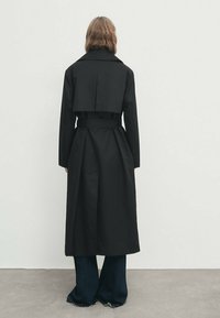 Musta trench-takki, jossa on kaksirivinen muotoilu, leveä kaulus, vyöllinen vyötärö ja keskipitkä leikkaus. Suunnikas, teksturoitu kangas.