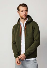 Felpa verde oliva con zip e finitura testurizzata, dettagli arancioni nella zip e tasca frontale, abbinata a una camicia bianca con colletto sotto.
