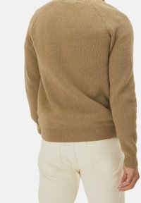 Beżowy sweter w prążki noszony przez osobę widoczną z tyłu, połączony z białymi spodniami na jednolitym tle.