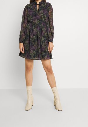 Kobieta w czarnej, kwiatowej mini sukience z długim rękawem, plisami i wiązaniem przy szyi, w połączeniu z kremowymi botkami na słupku.