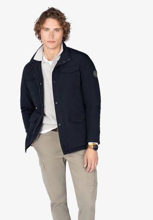 Hombre joven vistiendo una chaqueta azul marino sobre una camiseta polo gris claro y pantalones cargo beiges, de pie con una mano en el bolsillo contra un fondo blanco.
