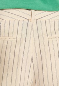 Béžové pruhované kalhoty s tmavými pruhy, vybavené upraveným pasem a dvěma zadními kapsami. Tkanina má hladkou, jemnou strukturu.