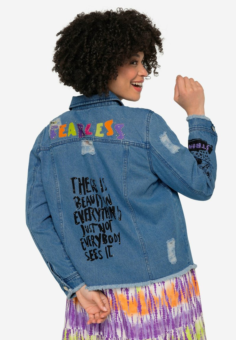 Jeansjacke, abgenutztes Design, blaue Farbe, mit mehrfarbigem "FEARLESS"-Text und aufgedrucktem Zitat auf der Rückseite. Ausgefranste Bündchen und Saum.