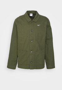 Vihreä puuvillajakku, jossa on kauluspaita, etupainikekiinnitys, kaksi suurta taskua ja pieni valkoinen Nike-logo vasemmassa rinnassa.