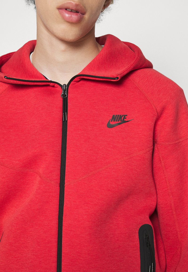 Czerwona bluza z zamkiem na suwak, wykonana z teksturowanej tkaniny, z czarnym logo Nike oraz boczną kieszenią zapinaną na suwak z czarnymi akcentami. Gładkie zapięcie na zamek.