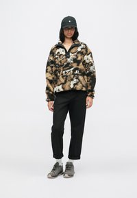 Pullover de fleece castanho e branco com padrão, gola alta, detalhe de botão preto e bolso frontal. Calças pretas e sapatilhas de caminhada cinzas.