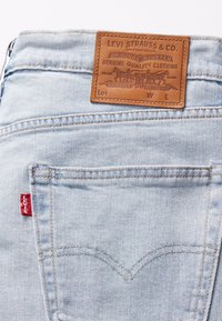 Lichtblauwe denim jeans hebben een leren label met ingedrukte tekst, een rode Levi's tab op de achterzak en zichtbare stikseldetails.