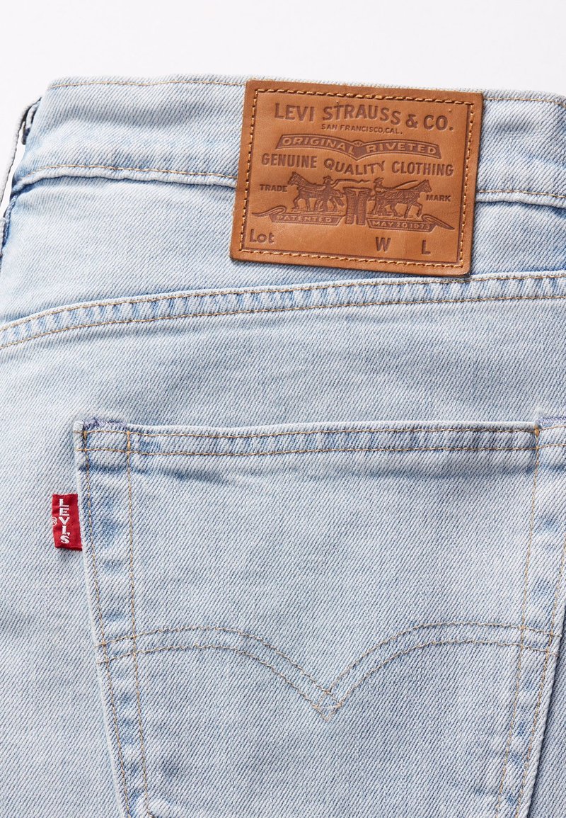 Un jean en denim bleu clair présente une étiquette en cuir avec un texte en relief, un onglet rouge Levi's sur la poche arrière et des détails de couture visibles.
