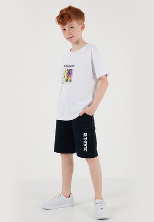 Rudy chłopiec ubrany w biały T-shirt z kolorowym wzorem i czarne szorty z napisem "AUTHENTIC", stojący z jedną ręką w kieszeni.