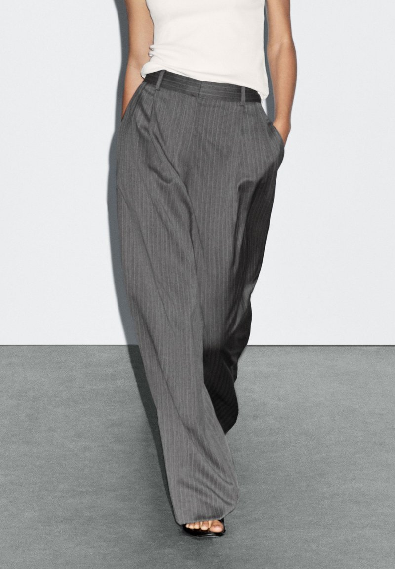 Gray pinstriped wide-leg trousers featuring front pleats and side pockets, made of a lightweight fabric, paired with a fitted white top.