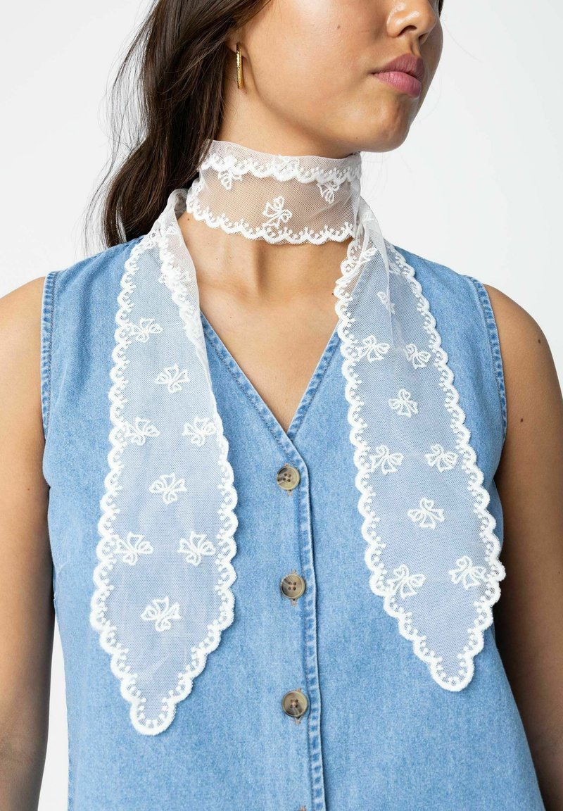 Écharpe en dentelle blanche avec broderie florale nouée autour du cou, portée sur un top bleu en denim sans manches à boutons, montrant le profil latéral d'une femme.