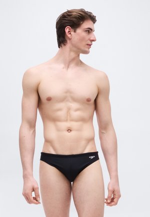 Fiatal férfi rövid barna hajjal, fekete Speedo úszónadrágot visel, és egy sima világosszürke háttér előtt áll, jobbra néz.