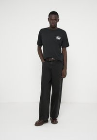 Μαύρο t-shirt με λευκό γραφικό, συνδυασμένο με φαρδιά μαύρα τζιν. Φοριέται με καφέ ζώνη και καφέ παπούτσια. Απλός σχεδιασμός, απλό φόντο.